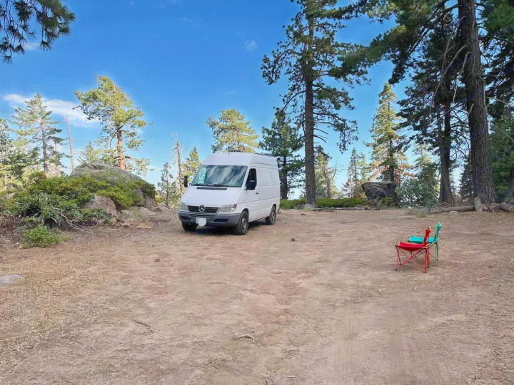 A white RV rental campervan is parked on a dirt clearing in a sunny forest campsite with two chairs.. Mercedes Sprinter 2006
