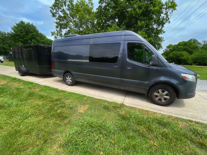 A grey RV rental Sprinter van towing a black trailer on a path beside green grass and trees.. Mercedes Sprinter Motorhome Campervan RV 2021