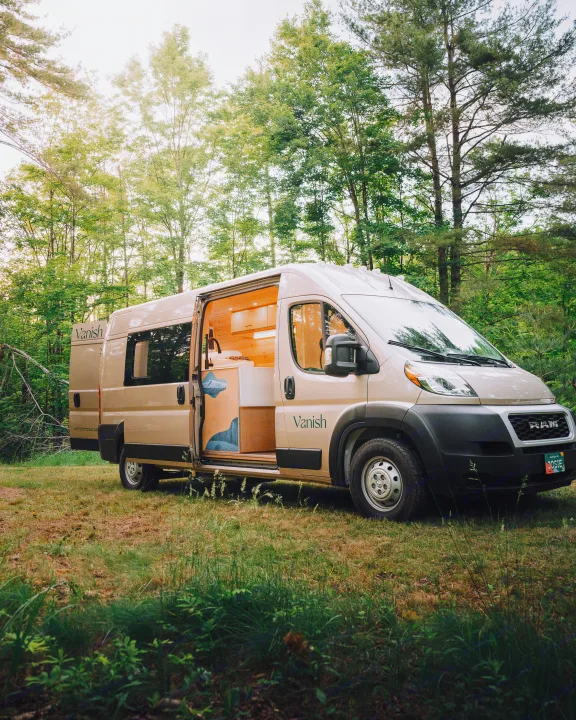 A beige Vanish camper van, ready for rv rental, rests in a lush forest, door open to reveal a warm, wood-paneled interior.. Ram Promaster 2021