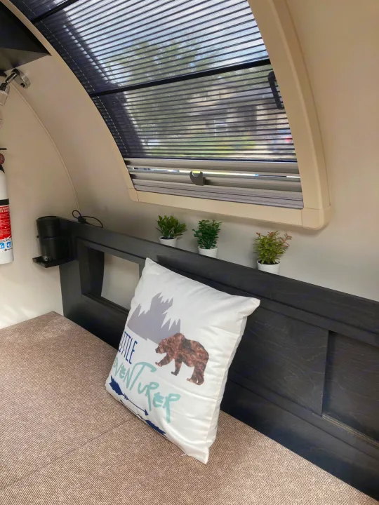 Inside a comfy rv rental: a bed with a bear pillow, plants, and a window with blinds, creating a cozy travel space.. Nucamp T@G 2022