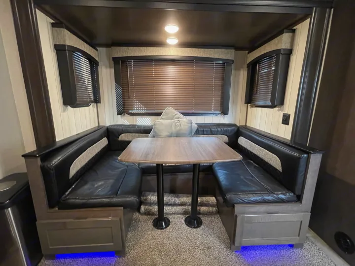 Spacious RV rental dinette featuring black booth seating, a wooden table, and blinds. Blue under-lighting illuminates the carpet.. Crossroads RV Sunset Trail Super Lite 2019