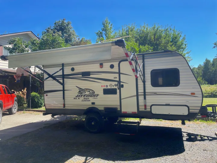 White Jay Flight RV rental with awning extended, parked on a sunny driveway. Red truck, house, and trees are in the background.. Jayco Jay Flight SLX 2016
