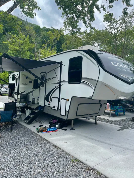 Modern fifth-wheel RV rental parked at a scenic campground. Awning extended, outdoor setup, with green mountains in the background.. Keystone RV Cougar 32BHS 2021