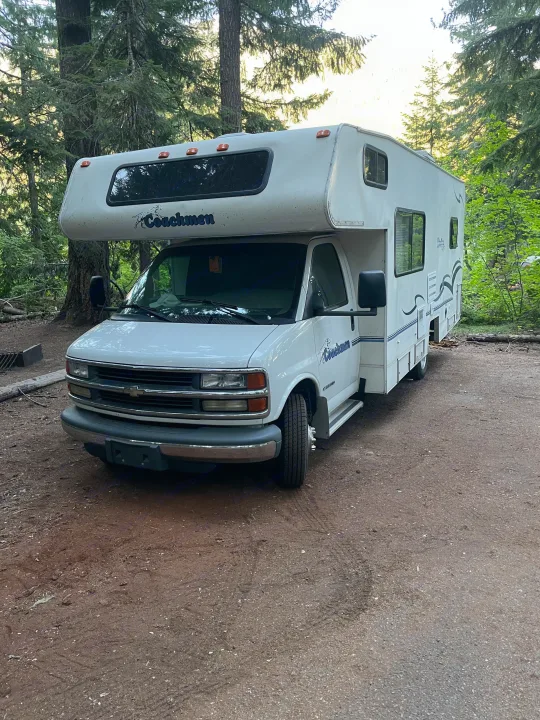 This white Coachmen rv rental is parked at a dirt campsite surrounded by towering trees, perfect for a forest escape.. Coachmen Catalina Sport 2002