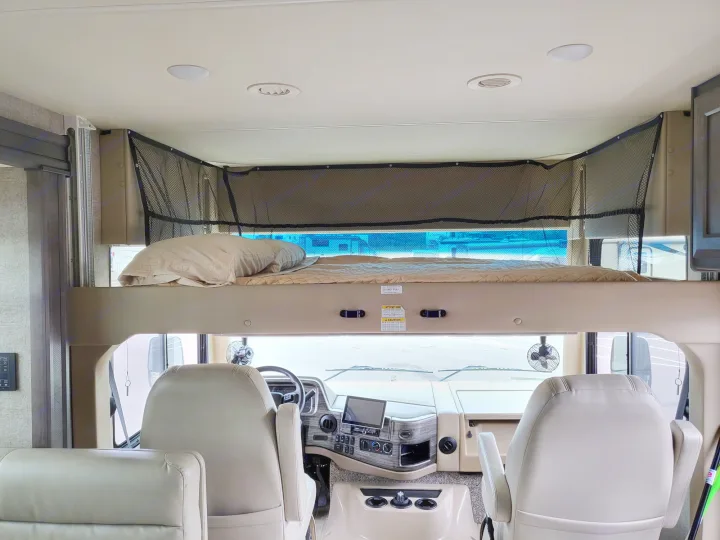 Inside an RV rental: front cabin with driver/passenger seats below a cozy overhead bunk bed, secured by a safety net.. Thor Miramar 2022