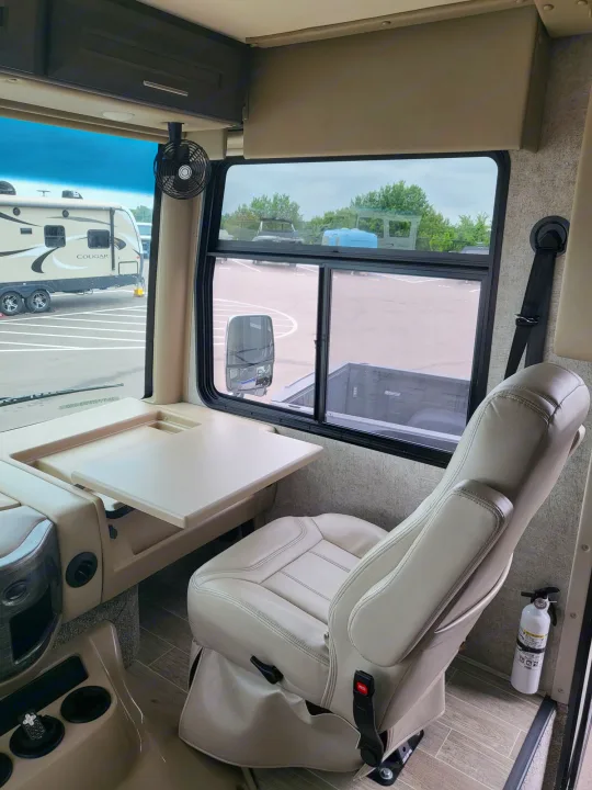 Interior of an rv rental, featuring a cream passenger swivel seat, pull-out table, and window views of other RVs and a truck. Fire extinguisher visible.. Thor Miramar 2022