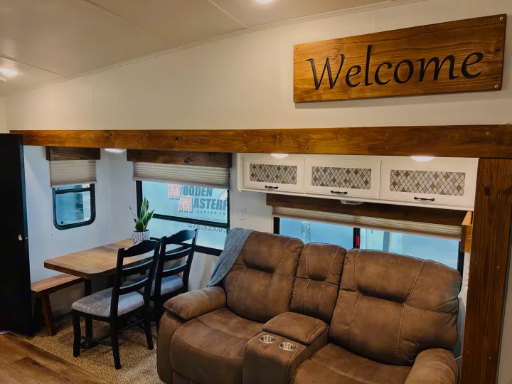 Spacious rv rental interior featuring a brown reclining sofa, wooden dining set, white cabinets, and a "Welcome" sign, with rustic wood accents.. Heartland RVs Greystone 2012