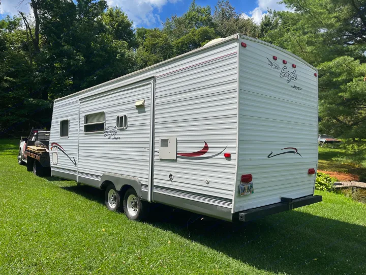 White Jayco Eagle travel trailer RV rental, parked outdoors on green grass and shaded by trees.. Jayco Eagle 2003