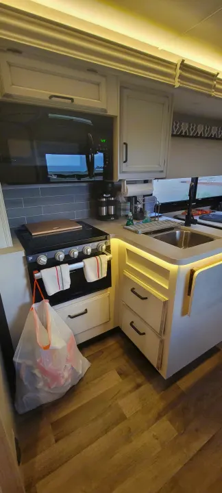 Modern RV rental kitchen with a microwave, stove, sink, and LED-lit counters. A trash bag hangs from the oven handle.. Jayco Greyhawk 2021
