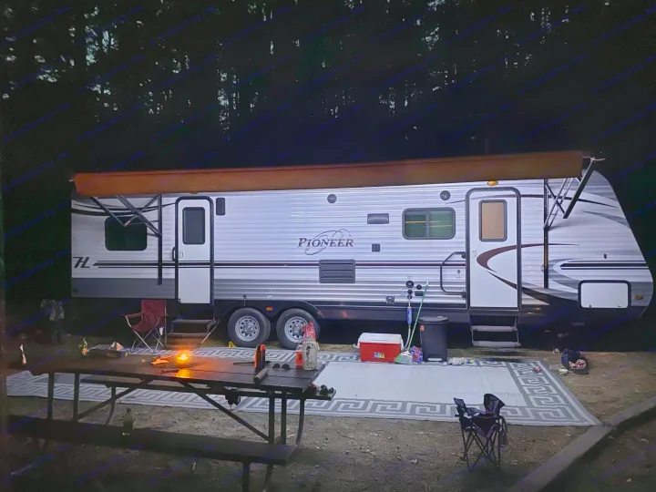 An RV rental parked at a lit campsite at night, with an outdoor rug and picnic table under surrounding trees.. Heartland RVs Pioneer Toy Hauler 2017