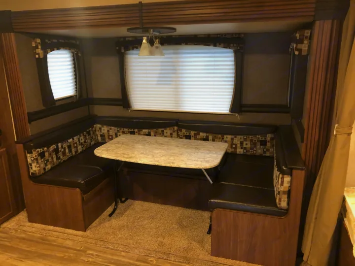 Inside an RV rental: a comfortable dinette with dark upholstered booth seating, a light table, and windows with blinds.. Heartland RVs Pioneer Toy Hauler 2017