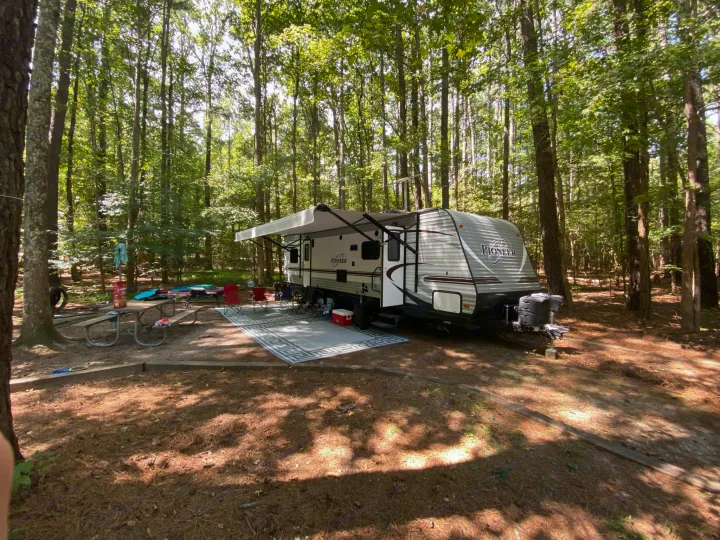 RV rental set up at a peaceful, sunny wooded campsite. A travel trailer with an extended awning, outdoor rug, and picnic table.. Heartland RVs Pioneer Toy Hauler 2017
