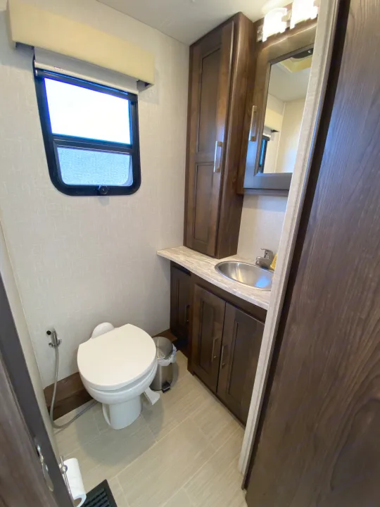 Compact RV rental bathroom: white toilet, dark wood vanity with sink & mirror, bright window, and tiled floor.. Coachmen Sports Coach 339DS 2019