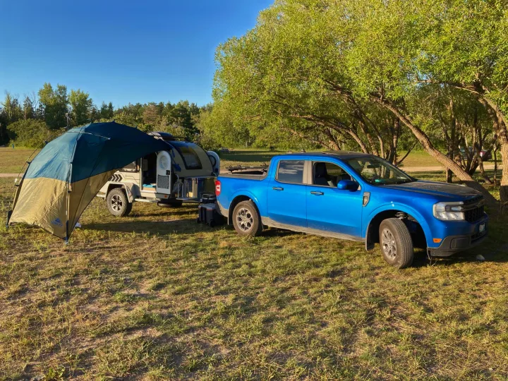 Sunny day setup: blue Ford Maverick towing a teardrop RV rental with an attached tent, parked on grass.. Nucamp T@G 2022