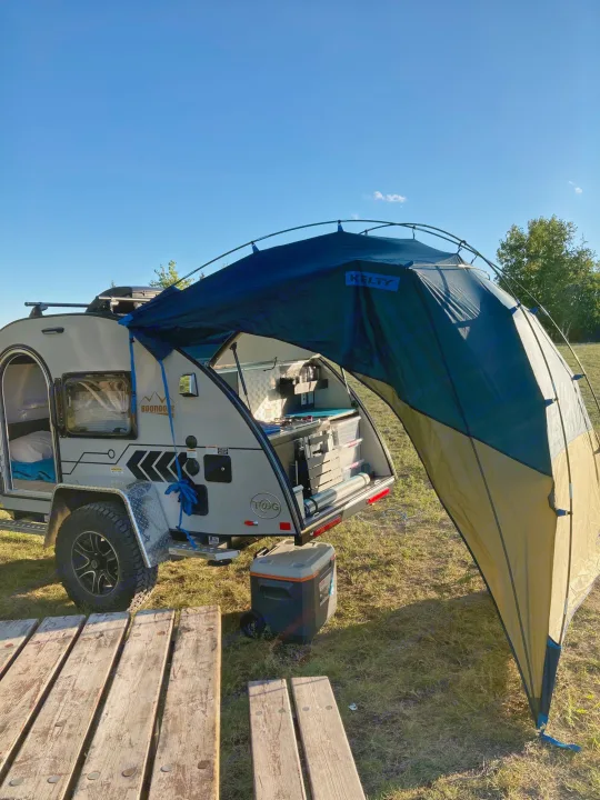 Teardrop RV rental camper with open rear kitchen and extended Kelty awning at a sunny campsite.. Nucamp T@G 2022