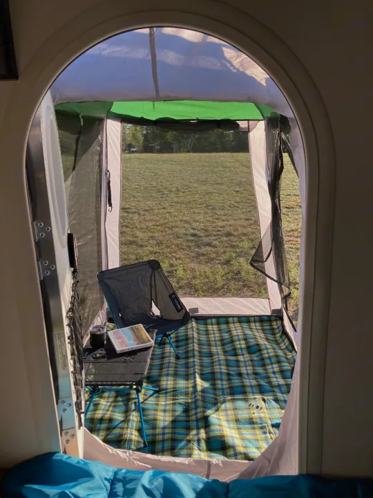 View from an rv rental, looking into an attached screen room setup with a folding chair, table, plaid mat, and grassy field.. Nucamp T@G 2022