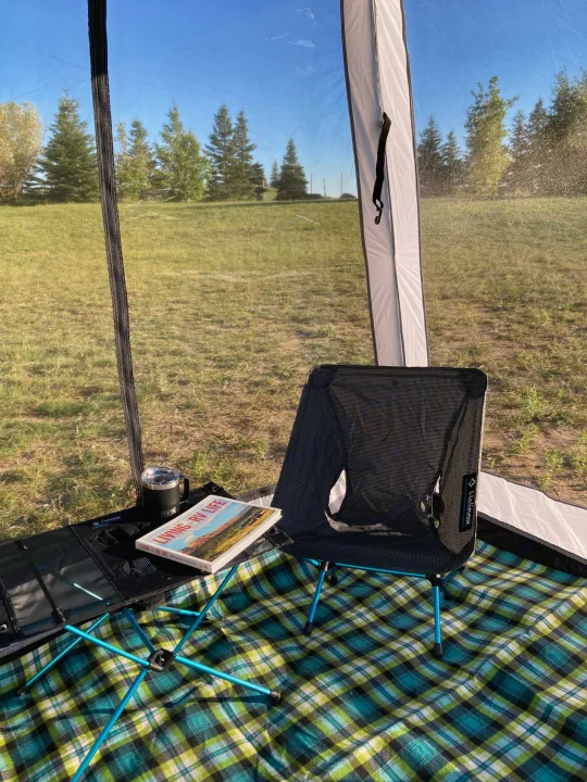 Cozy RV rental setup: Helinox gear, Yeti mug, and "Living RV Life" book inside a screen room, overlooking a sunny field.. Nucamp T@G 2022