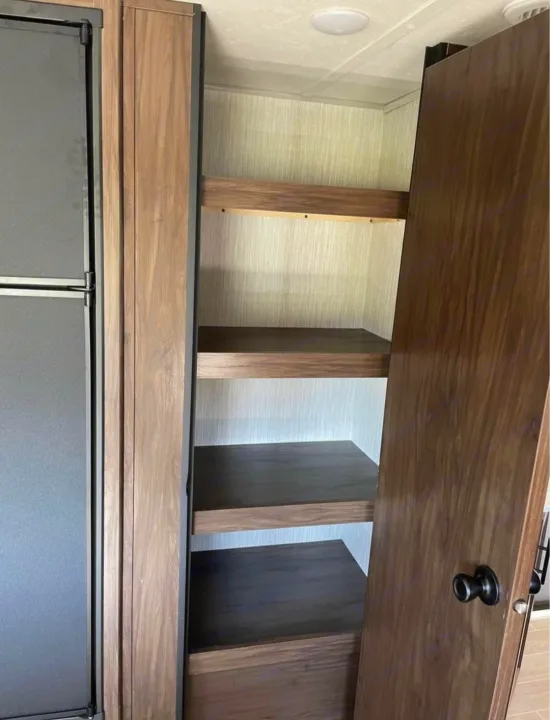 Organized RV rental pantry: wood shelves, textured wall, and a dark refrigerator. Perfect for travel storage.. Forest River Cherokee Grey Wolf 2021
