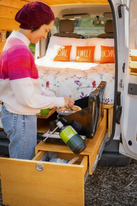 An RV rental setup for van life: woman cooking on a pull-out stove outside a camper van with a cozy bed.. Nissan NV200 2019