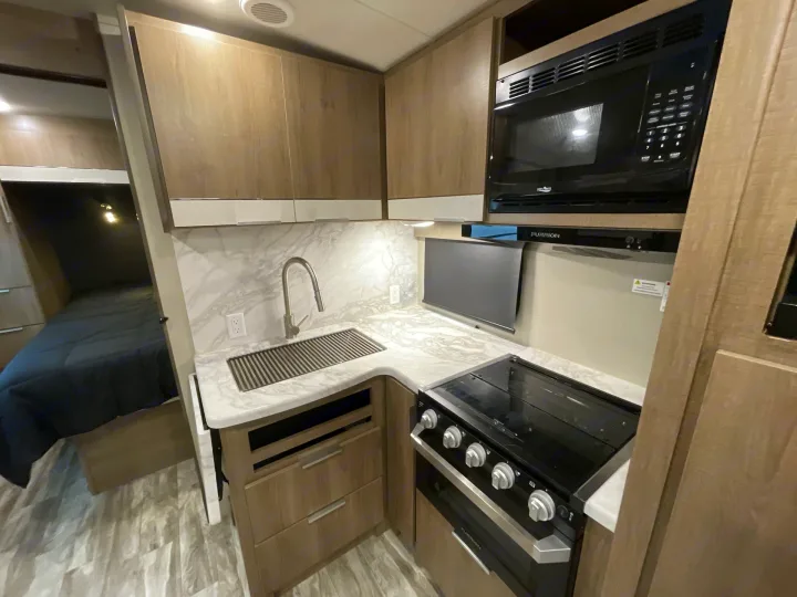Modern RV rental interior featuring a compact kitchen with stove, microwave, sink, and adjacent bedroom.. Grand Design Imagine 2400BH 2021