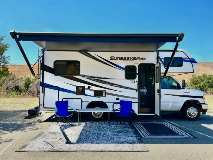 A white Sunseeker RV rental with an extended awning, chairs, and rugs set up for a cozy outdoor campsite experience.. Forest River Sunseeker 2022