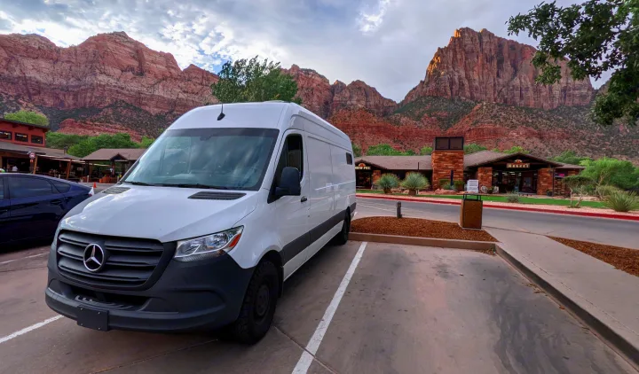 White Mercedes Sprinter RV rental parked in Zion National Park, with towering red mountains and village buildings in the background.. Mercedes-Benz Sprinter 2020