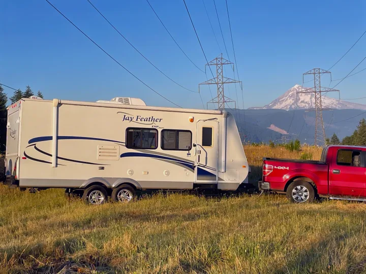 Your RV rental journey begins! A red pickup tows a Jay Feather travel trailer in a sunny field with a majestic mountain and power lines.. Jayco Jay Feather Exp 2010