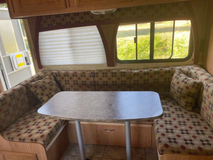 Inside an RV rental: a spacious dinette with patterned U-shaped seating, center table, and large windows revealing green outdoor scenery.. Jayco Jay Feather Exp 2010