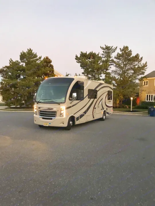 Beige Thor Vegas RV rental on a street at dusk, headlights on, with trees and houses in background. Perfect for travel.. Thor Vegas 2016