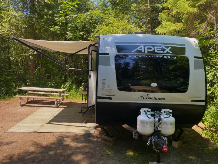 This RV rental is set up at a wooded campsite, featuring an extended awning, outdoor mat, and picnic table for a comfortable stay.. Coachmen Apex 2021