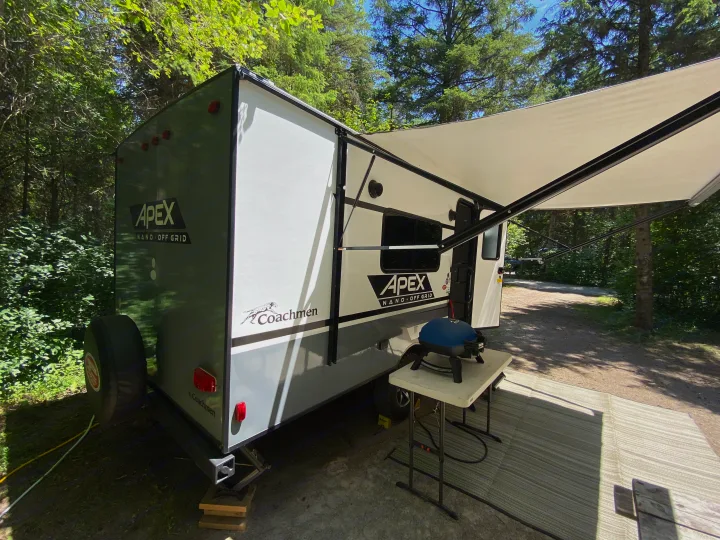 An RV rental set up for camping: a travel trailer with an extended awning, outdoor mat, and a grill on a table.. Coachmen Apex 2021