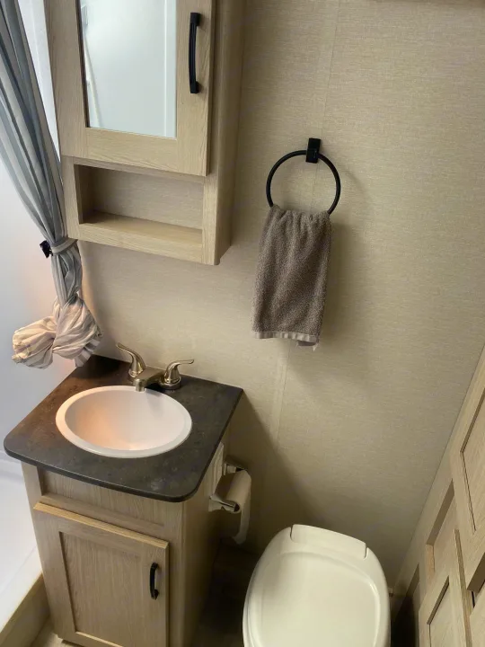 Inside an RV rental: a compact bathroom with light wood cabinets, sink, toilet, and striped shower curtain.. Coachmen Apex 2021