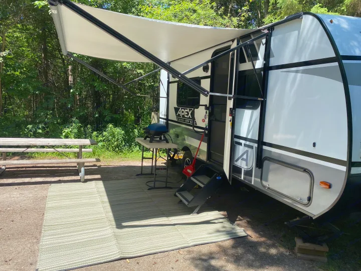 This RV rental camping setup features an extended awning, picnic table, grill, and outdoor mat in a treed campsite.. Coachmen Apex 2021