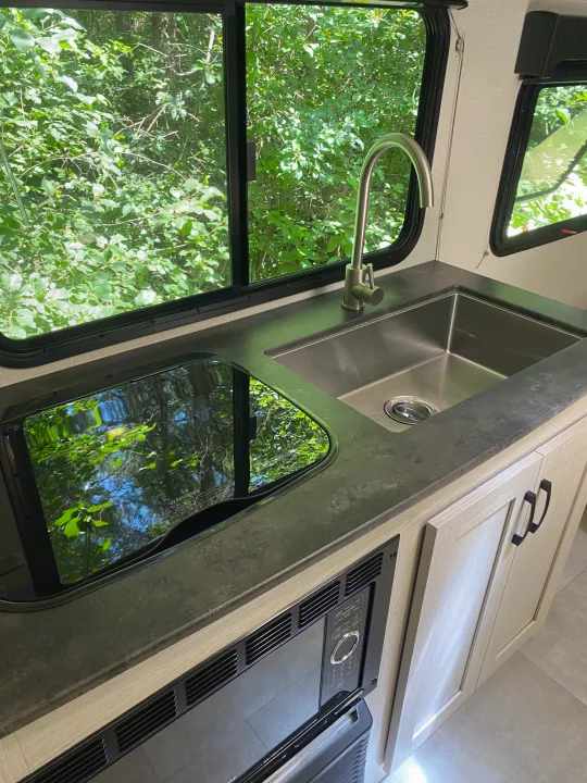 RV rental kitchen featuring a stainless steel sink, faucet, covered cooktop, and microwave, with lush green views outside.. Coachmen Apex 2021