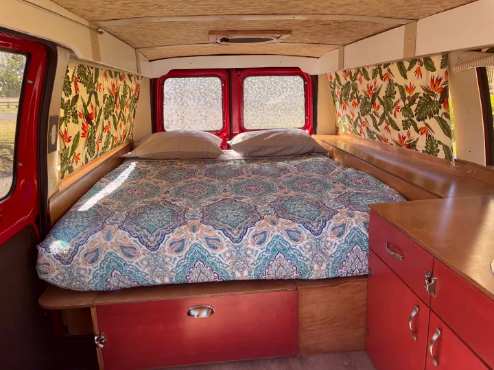 Inside this cozy RV rental: a comfortable bed with tropical print curtains and red cabinets, ideal for travel.. Ford Ford E350 2010