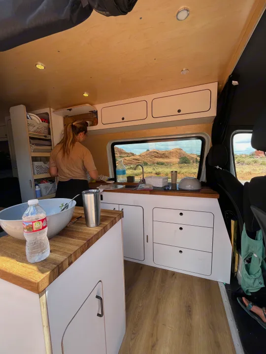 Woman cooking in a well-equipped RV rental kitchen. Camper van interior with wood countertops & a scenic desert view.. Mercedes-Benz Sprinter 419 LWB High Roof 2022