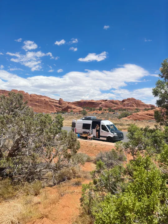 RV rental parked on desert road by red rock cliffs. Two people relax outside white camper van under blue, cloudy sky.. Mercedes-Benz Sprinter 419 LWB High Roof 2022