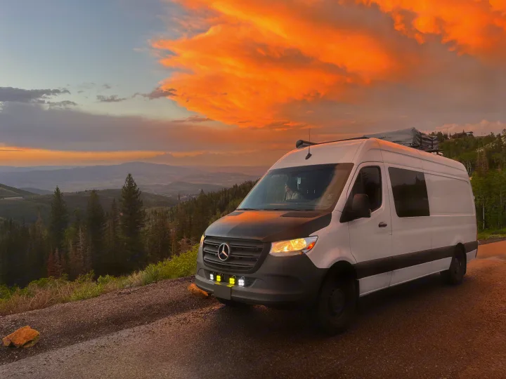 A white RV rental van overlooking a sunset-lit valley. Vibrant orange clouds, mountains, and forest. Ready for adventure.. Mercedes-Benz Sprinter 419 LWB High Roof 2022