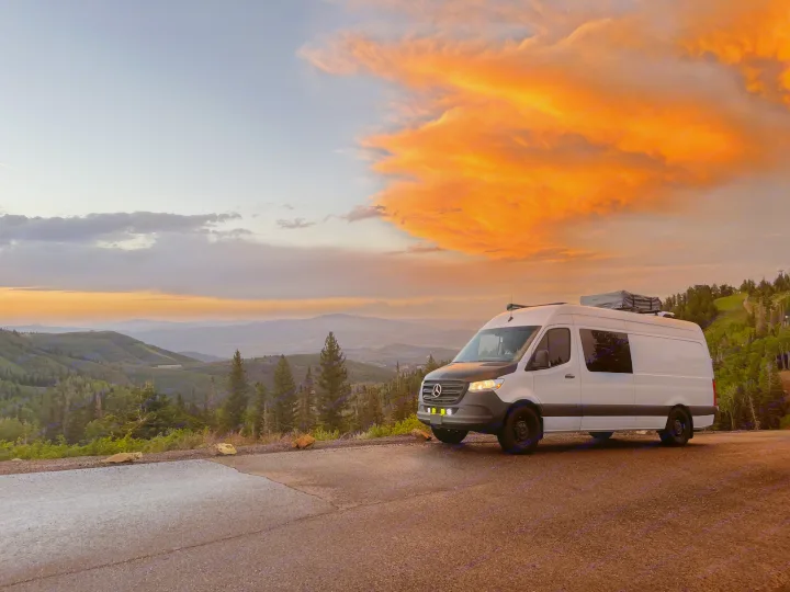 White Mercedes RV rental van at a mountain overlook. Vibrant orange sunset clouds fill the sky above scenic valleys.. Mercedes-Benz Sprinter 419 LWB High Roof 2022