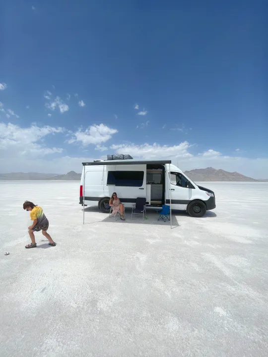 RV rental on vast salt flats under a blue sky. A woman relaxes under the awning, a child plays nearby. Road trip fun!. Mercedes-Benz Sprinter 419 LWB High Roof 2022