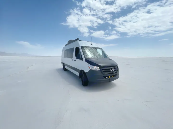 A white and black RV rental van parked on a vast salt flat, with distant mountains and a blue sky overhead.. Mercedes-Benz Sprinter 419 LWB High Roof 2022