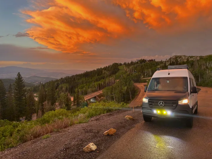 RV rental van on a mountain road at sunset, with glowing orange clouds over forested peaks. Scenic drive, perfect for outdoor adventure.. Mercedes-Benz Sprinter 419 LWB High Roof 2022