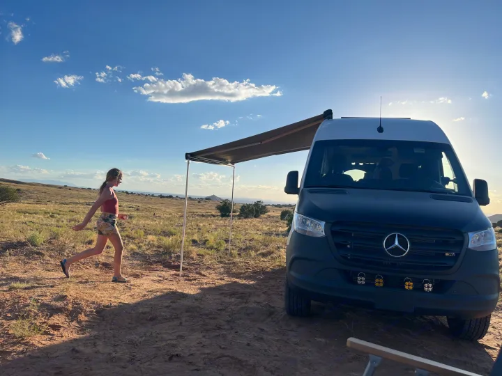 RV rental adventure: A woman walks by a camper van with an awning in a sunny, expansive desert landscape.. Mercedes-Benz Sprinter 419 LWB High Roof 2022