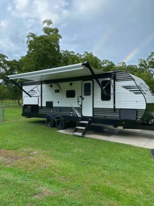 Experience camping with an RV rental! This white travel trailer with an extended awning is parked by grass under a double rainbow sky.. Highland Ridge RV Olympia 2022