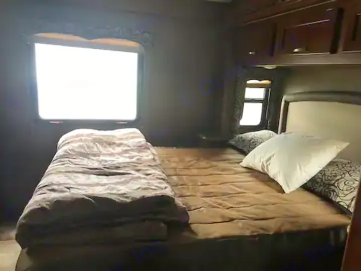 Cozy RV rental bedroom with a queen bed, brown bedding, two bright windows, and overhead cabinets.. Thor Four Winds 2017
