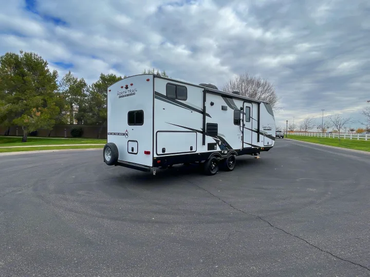 A large white travel trailer, a great option for an RV rental, parked outdoors on asphalt with trees and a cloudy sky.. Heartland North Trail 2022