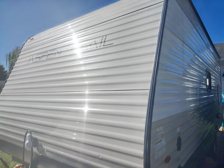 The corrugated light grey side of an Aspen Trail RV rental shines in direct sunlight, revealing its front angled view.. Dutchmen 17BH 2021