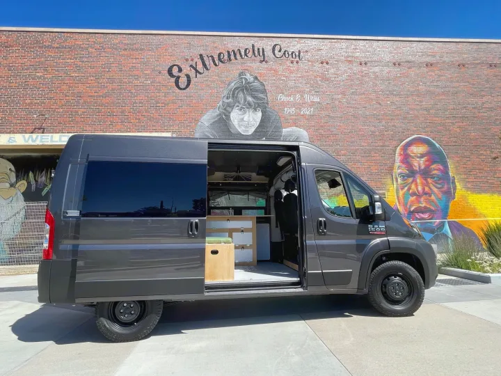 Custom RV rental: A grey camper van with open door showing wood interior, parked by vibrant Chuck E. Weiss wall mural.. Ram Promaster 2022