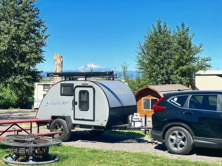 Bushwhacker teardrop camper, hitched to an SUV, at an RV rental site with a snow-capped mountain backdrop.. Braxton Creek Bushwhacker 2021