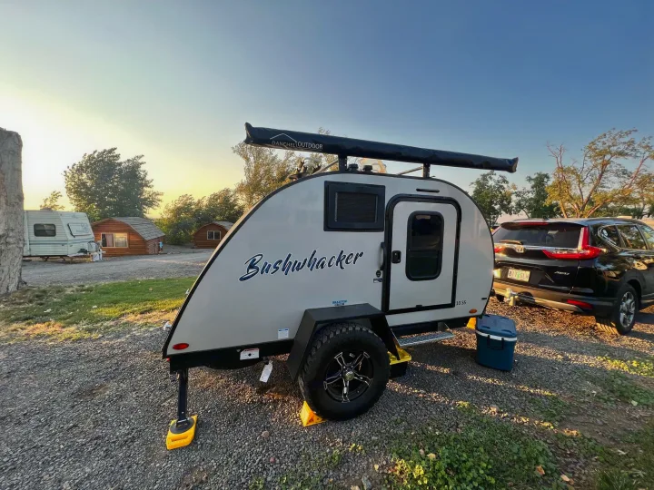 A Bushwhacker 10 SS teardrop RV rental, hitched to an SUV and with a cooler, parked on gravel for outdoor travel.. Braxton Creek Bushwhacker 2021